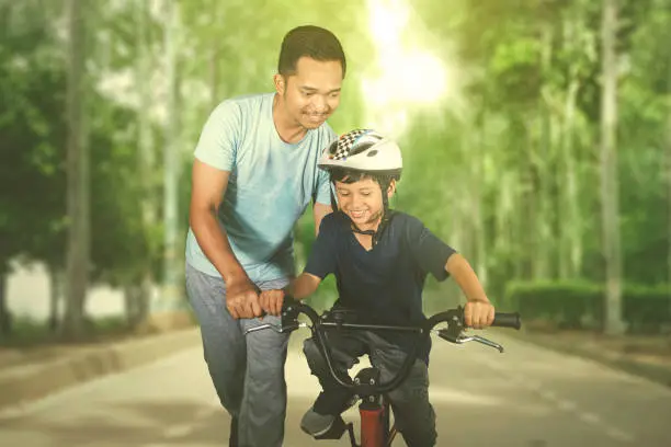 Picture of a happy little boy learning to ride a bicycle with his father in the park. Shot at summer time