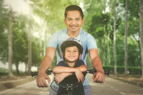 Picture of Asian man looking at the camera while riding bicycle with his son in the park