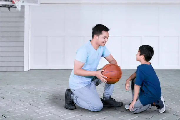 Picture of young man kneeling in the garage while teaching his son to play basketball