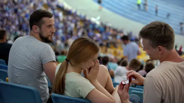 Guy making proposal to girlfriend at stadium during football match, surprise