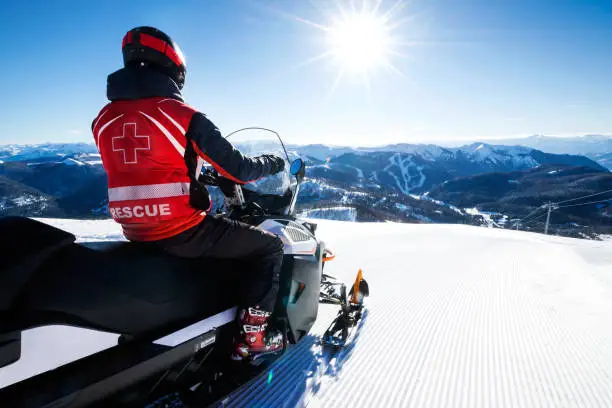 Photo of Search and rescue worker sitting on his motor sleds, looking at a beautiful mountainside view on a bright, sunny day