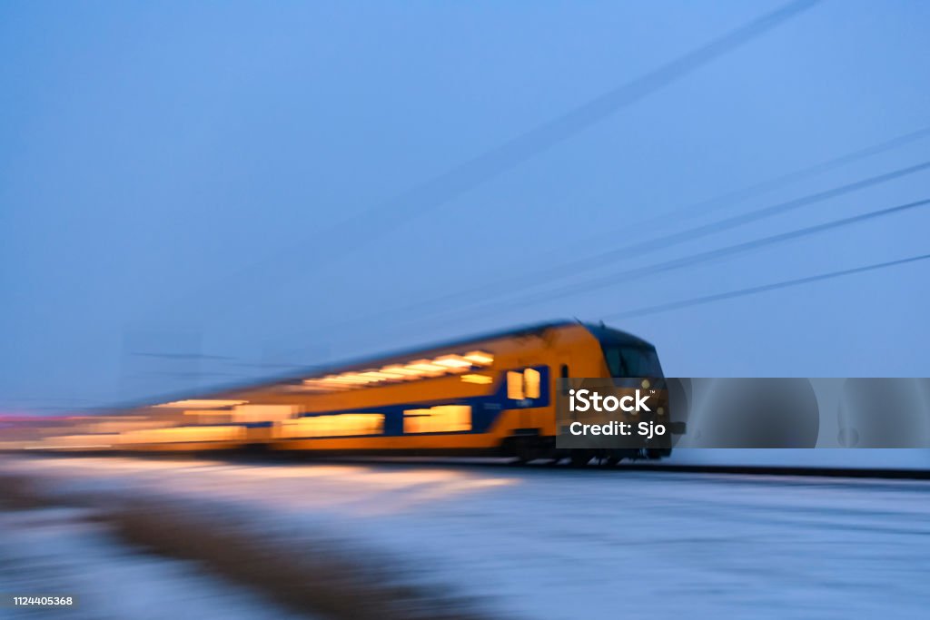 Intercity train of the Nederlandse Spoorwegen (NS) driving through the snow during a cold winter evening Blue and yellow Dutch intercity train of the Nederlandse Spoorwegen (NS) driving through the snow on a cold winter day. Snow is blowing up from the ground as the train is passing. Night Stock Photo