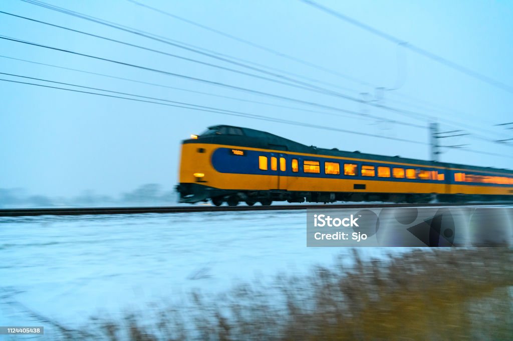 Intercity train of the Nederlandse Spoorwegen (NS) driving through the snow during a cold winter evening Blue and yellow Dutch intercity train of the Nederlandse Spoorwegen (NS) driving through the snow on a cold winter day. Snow is blowing up from the ground as the train is passing. Blue Stock Photo