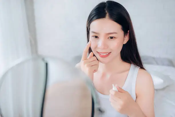 Beauty Asian Woman use Acne Gel Skincare after Cleansing on Face His Happiness and Look at mirror in White Bedroom.