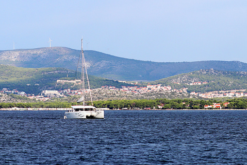 Sailing catamaran sails along the rocky green coast past the red literary sign - the fairway lighthouse. City of Sibenik in the Dalmatia region in Croatia. Adriatic sea in the Mediterranean area.
