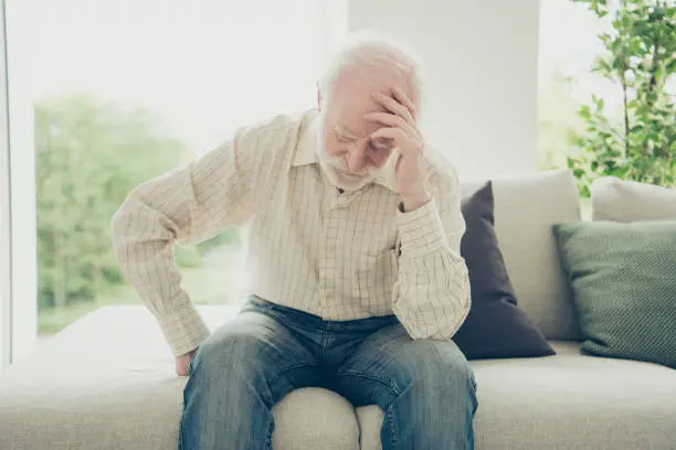 Close up portrait of grey haired he his him grandpa with chronic strong terrible headache head on hand illness wearing casual checkered shirt jeans denim outfit sitting comfy on divan