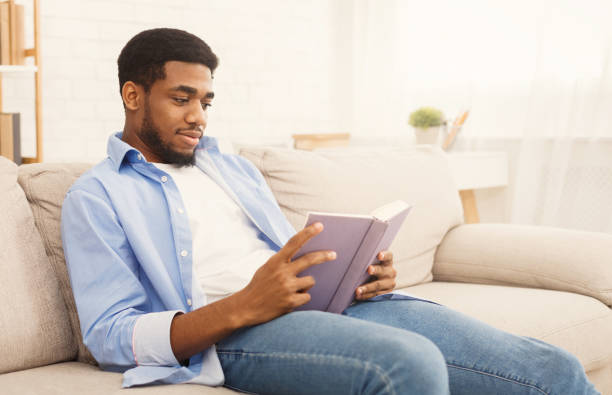 istock Young african-american man reading book at home 1124421162