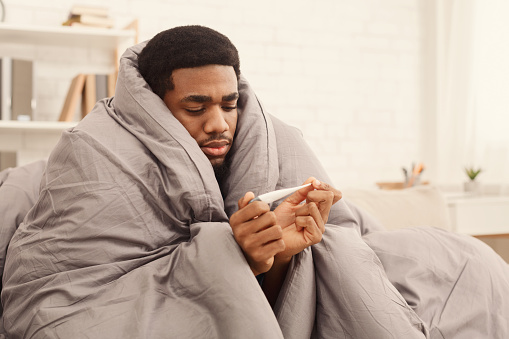 Ill african-american man covered with blanket holding thermometer, having fever, copy space