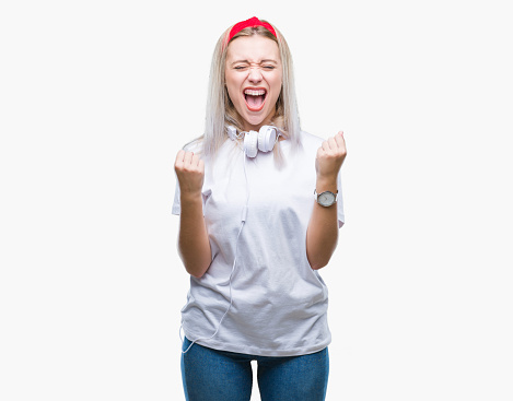 Young blonde woman wearing headphones listening to music over isolated background celebrating surprised and amazed for success with arms raised and open eyes. Winner concept.