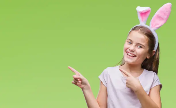 Photo of Young beautiful girl wearing easter bunny ears over isolated background smiling and looking at the camera pointing with two hands and fingers to the side.