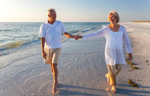 Happy senior man and woman couple walking and holding hands on a deserted tropical beach with bright clear blue sky stock photo