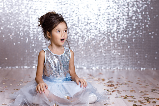 little child girl in blue dress sitting on the floor with confetti on background with silver bokeh. birtday party