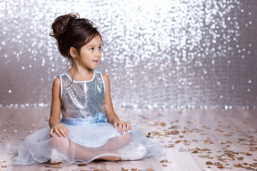 little child girl in blue dress sitting on the floor with confetti on background with silver bokeh. birtday party