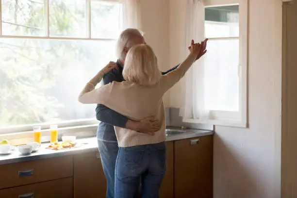 Active aged grey haired couple husband and wife wearing casual clothes after cooking healthy breakfast enjoying time embracing dancing in modern light kitchen in the morning, female rear back view