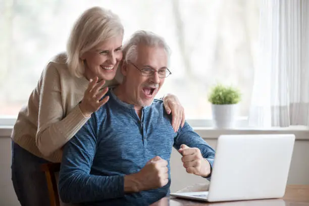 Happy aged spouses looking on pc screen feels happy received unbelievable great news, enjoying celebrating win online lottery. Senior positive wife husband founding out about sale and huge discounts