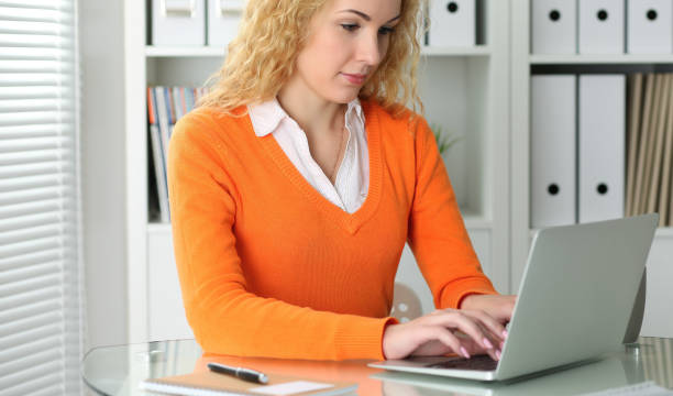 istock Young beautiful business woman or student communicates by laptop computer while sitting at the desk and holding black pen. She Orange sweater is at the girl. Education, business and lifestyle concept. 1124441170