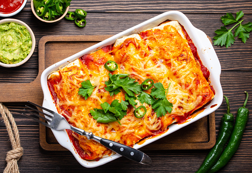 Traditional Mexican dish enchiladas with meat, chili red sauce and cheese in white casserole dish over rustic wooden background, served with guacamole and tomato dips. Close up, top view