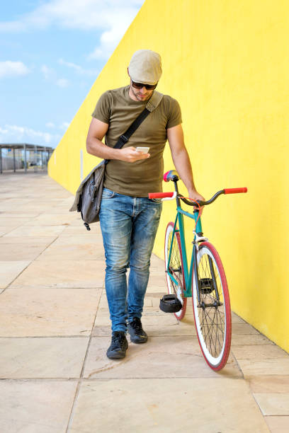 istock Front view of a young trendy man with a fixed bike wearing casual clothes while using a mobile phone against a yellow wall outdoors in a sunny day 1124451600