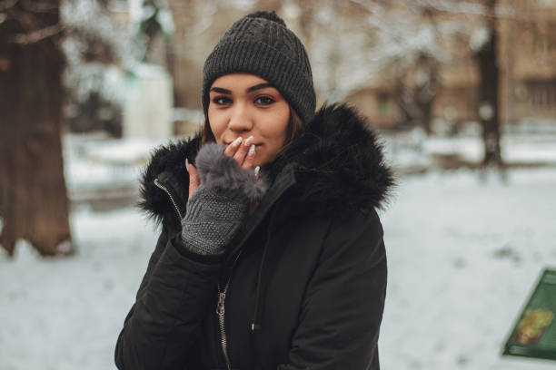Beautiful young woman in winter clothes standing in park covered with snow and sending a kiss.