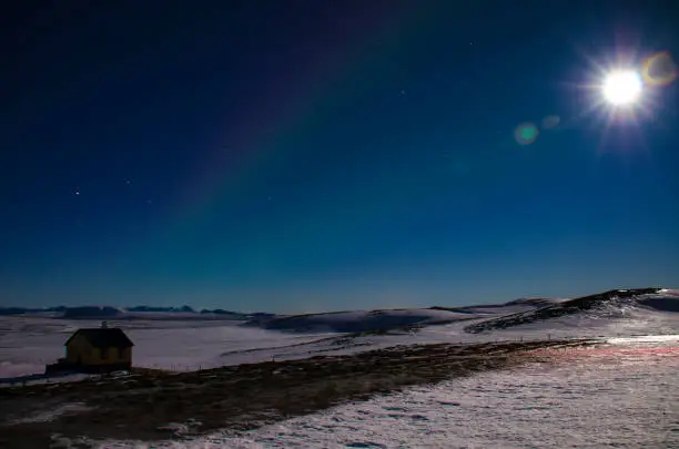 The moon with the rays and the Aurora in Iceland s blue winter sky over an Icelandic house that stands on a lava field covered with snow.