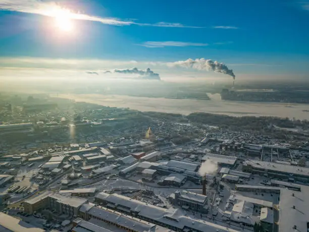 Photo of Winter landscape from a aerial view of the city of Novosibirsk in the haze with streets, small buildings, river under ice covered by snow and protruding pipe from which smoke comes