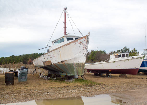 Old boat in marina shipyeard awaiting repairs against a cloudy sky. You can see white and green paint on the hull of the ship, the cabin exterior has rust stains on it. This old fashioned wooden motorboat is in a state of disrepair. Taken in a marina and dock in on the Chesapeake bay in coastal virginia.