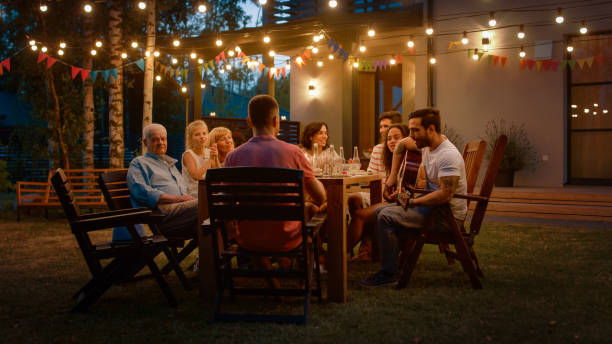 sitzen an den esstisch hübscher junger mann spielt gitarre für eine freunde. familie und freunde anhören von musik bei der sommergarten abend party feier. - family summer portrait nature stock-fotos und bilder