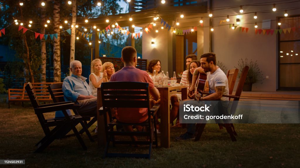 Assis à la Table du dîner beau jeune homme joue de la guitare pour un amis. Famille et amis, écouter de la musique lors de la célébration de fête de jardin de soirée d’été. - Photo de Famille libre de droits