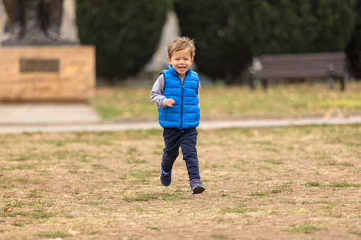 Cute boy in a blue vest smiling and running in the park. Selective focus.