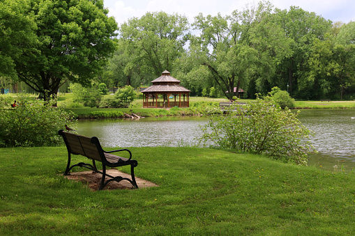 Beautiful late spring landscape with bench in a foreground, trees around the pond and wooden gazebo in a city park. Lakeview park, Middleton, Madison area, WI, USA.
