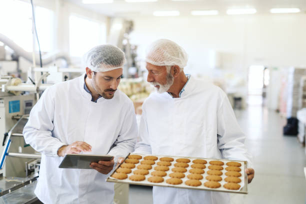istock Worker holding casserole with cookies while supervisor checking quality and holding tablet. Food factory interior. 1124533189
