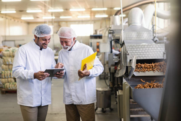 dos trabajadores uniformes mirando tablet estando en fábrica del alimento. mayores uno apuntando a la tableta. - sales manager fotografías e imágenes de stock