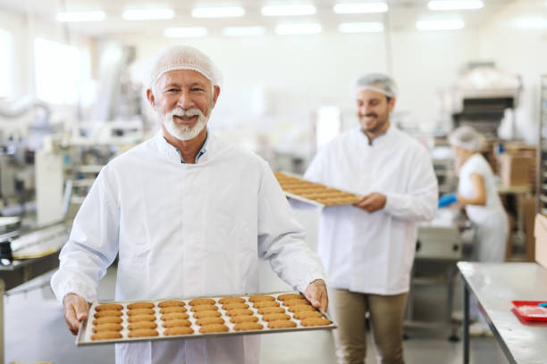 dos sonrientes trabajadores uniformes estériles llevan guisados con galletas. interior de la fábrica de alimentos. - quick cookies fotografías e imágenes de stock