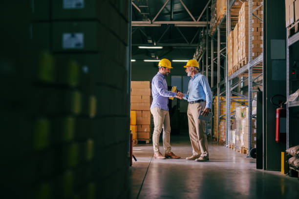 dos hombre de negocios con cascos de cabeza agitando las manos para bien terminado. interior de almacenamiento. - manual worker handshake industry warehouse fotografías e imágenes de stock