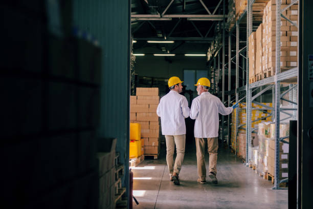 Two storage workers in uniforms and helmets on heads walking in storage and talking. Backs turned.