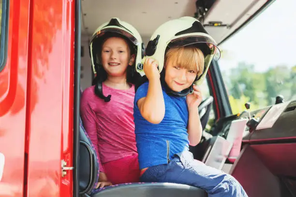 Two cute kids playing in fire truck, pretending to be firefighters, open doors day at fire station. Future profession for children. Educational program for schoolkids