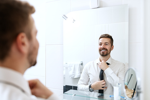 Sonriente hombre de negocios poniendo de corbata al mirarse en el espejo y de pie en el cuarto de baño. photo