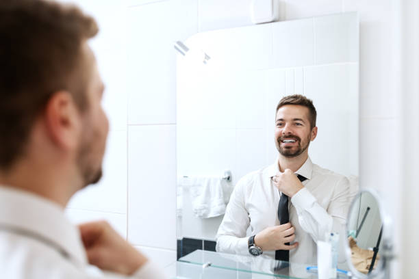 Smiling businessman putting on necktie while looking in the mirror and standing in the bathroom. Smiling businessman putting on necktie while looking in the mirror and standing in the bathroom. man adjusting tie stock pictures, royalty-free photos & images