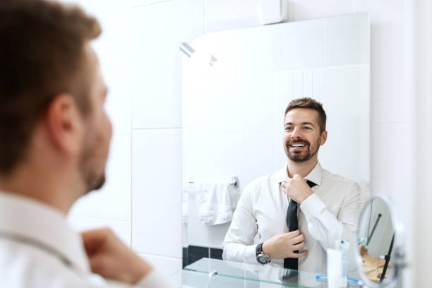 smiling businessman putting on necktie while looking in the mirror and standing in the bathroom. - shirt necktie men businessman imagens e fotografias de stock