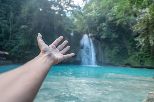 Personal perspective of female stretching out hand to a beautiful waterfall located on Cebu Island in the Visayas Islands in the Philippines. People travel sharing environment conservation concept.