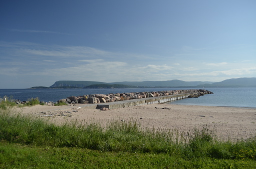 Man made seawall at Pleasant Bay Nova Scotia Canada, with sandy beach, grass in foreground and rolling hills across the water in the background.