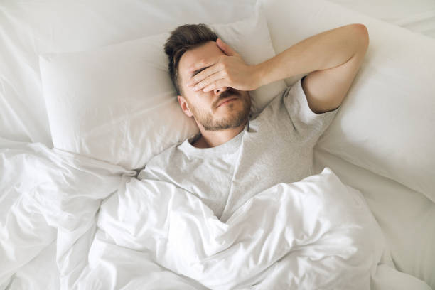 hombre joven cansado en la cama cerrando los ojos antes de despertar. mañana de resaca. trastorno del sueño y problemas. - tired man fotografías e imágenes de stock