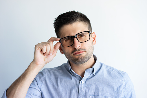 Pensive guy in casual touching temple of eyeglasses. Closeup of young Caucasian man adjusting glasses and staring at camera. Sight concept