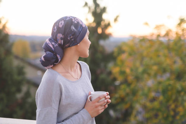 woman battling cancer standing outside and looking toward the mountains A beautiful young woman wearing a head wrap looks off in the distance and smiles contemplatively while drinking a cup of tea. She is standing outdoors and there are mountains and trees in the background. cancer cell photos stock pictures, royalty-free photos & images