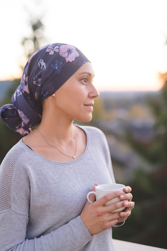 A beautiful young woman wearing a head wrap looks off in the distance and smiles contemplatively while drinking a cup of tea. She is standing outdoors and there are mountains and trees in the background.
