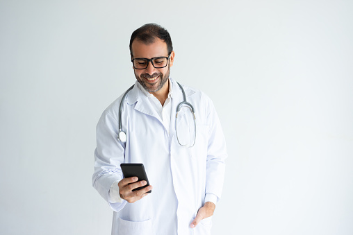 Smiling handsome male doctor reading news on smartphone. Middle-aged guy wearing white coat. Technologies in medicine concept. Isolated front view on white background.