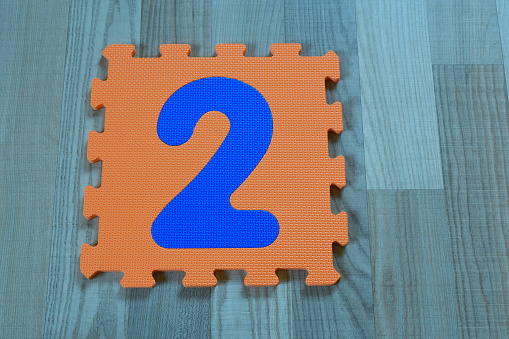 Orange puzzle piece with blue number two on wooden texture laminate floor indoors. Second birthday, anniversary and educational children toys for learning shapes, colors and numbers concept.