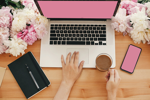 Girl hands on stylish laptop with empty screen and coffee, phone, black notebook and peonies on rustic wooden table. Freelance concept Flat lay. Workplace. Space for text.  Women's Day