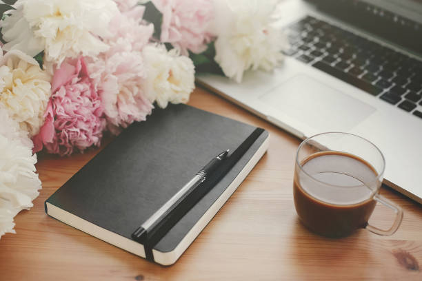 Stylish laptop, phone with empty screen, black notebook, coffee and peonies on rustic wooden table. Freelance concept. Woman workplace. Space for text. International Women's Day.