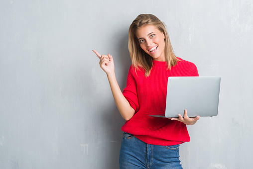 Beautiful young woman over grunge grey wall using computer laptop very happy pointing with hand and finger to the side